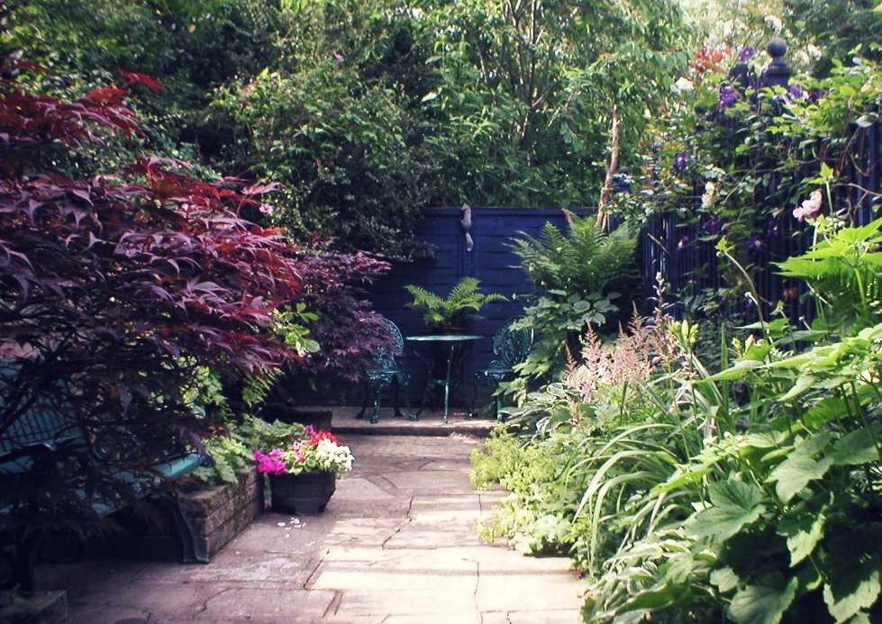 a paved path through overflowing flower beds, leading to a table and chairs at the end of the garden