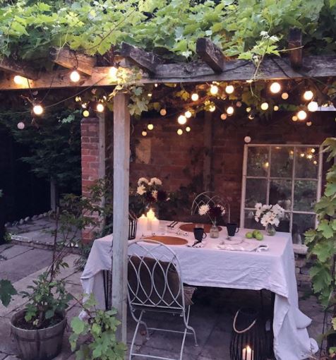 a dining area with pretty white tablecloth under a pergola covered with string lights and leafy vines