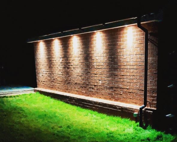 the side of a garage with bright downlighters illuminating the grass below