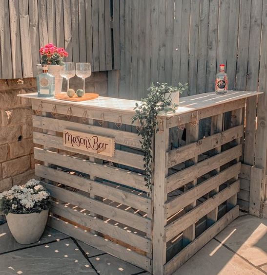 A pallet bar in the corner of a patio, in the same grey as the garden fence. Several potted plants decorate the counter and surrounding floor.