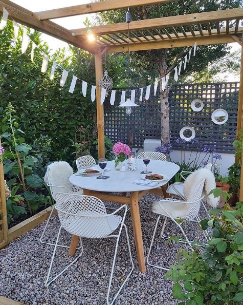 A slender-framed set of dining table and chairs on a gravel patio, under a pergola with festoon lights
