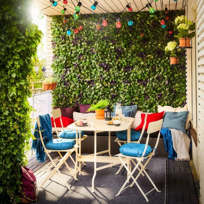 Gravity-Defying Green Wall Ideas 3 A small balcony with folding chairs and table, coloured string lights and artificial plant tiles covering the walls