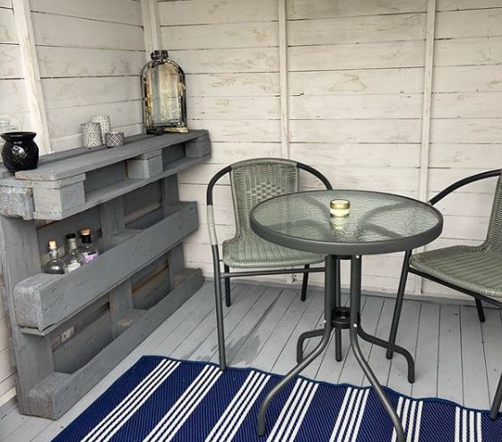 A small shed with table, chairs and a shelf made from a wooden pallet, housing some bottles.
