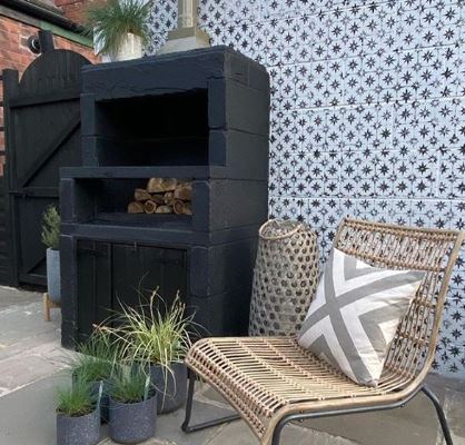A stylish wood-fired pizza oven surrounded by a wicker chair and pot plants on a patio