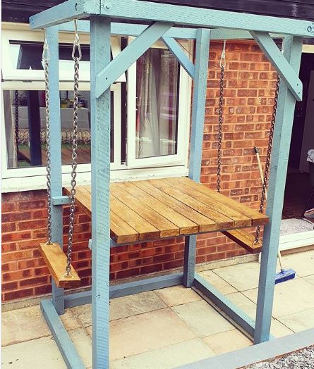 A blue wooden frame supports two narrow wooden swings facing a small dining table