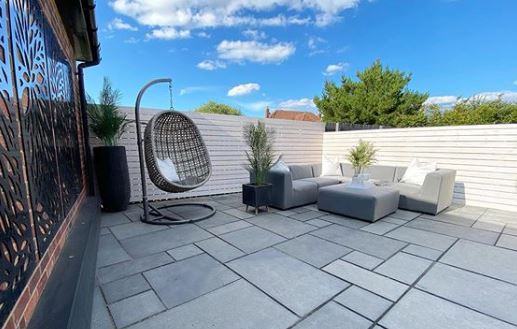 A grey swing chair sits in a minimalist garden with grey flagstones, white fence and grey garden sofas