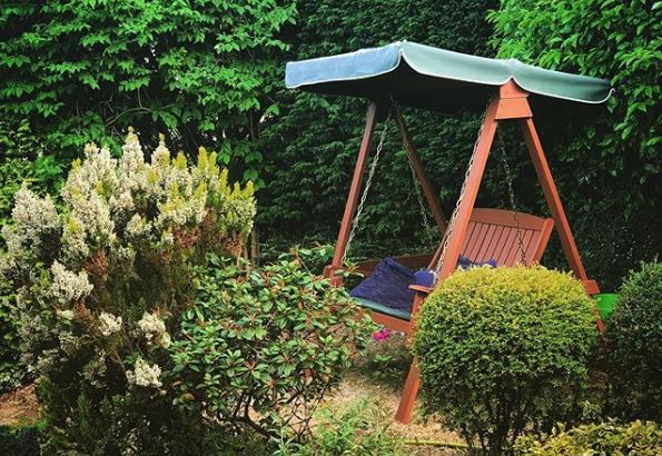 A traditional teak garden swing bench with canopy is tucked between hedges and shrubs
