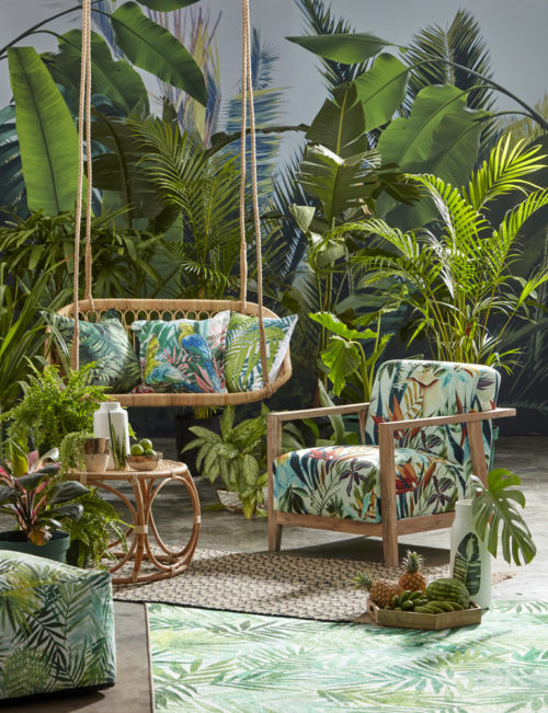 a swing bench and armchair in a tropical garden, both with cushions in a lurid leafy print