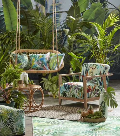 a swing bench and armchair in a tropical garden, both with cushions in a lurid leafy print