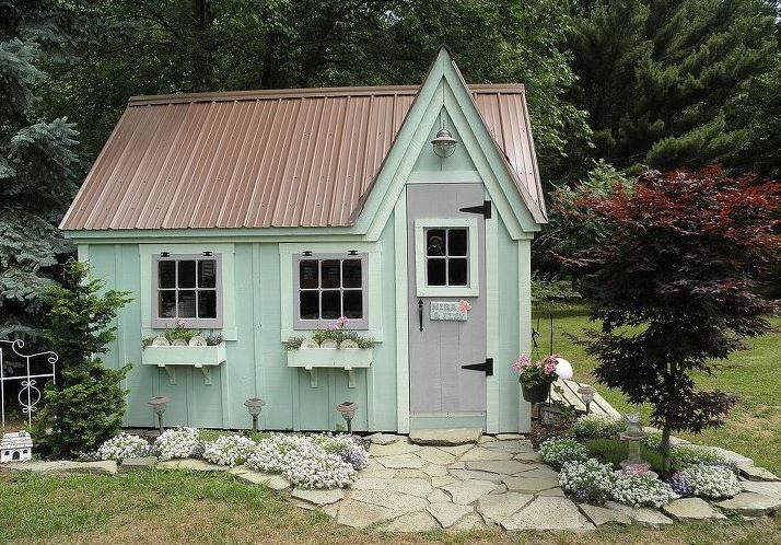 The exterior of a quaint garden shed, painted pale green, with a pitched roof and window boxes filled with flowers