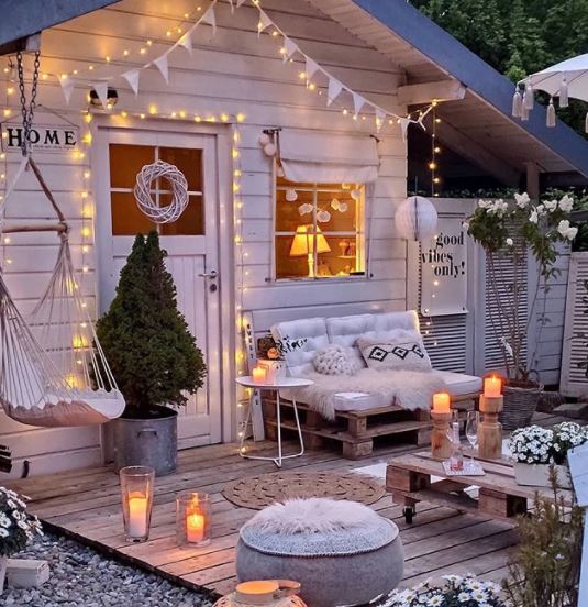 a busy deck outside a she shed, with pallet chair, pouffe, swing chair, bunting and candles, all in shades of cream and pale grey