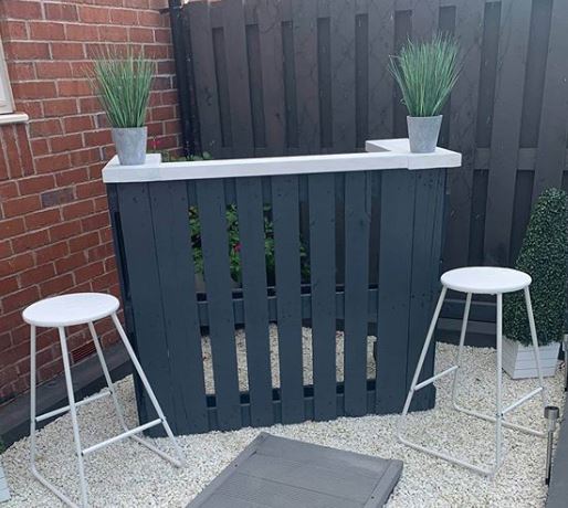 a dark grey pallet bar with white countertop and white stools, on a pale gravel patio