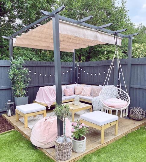 a small deck covered by a pergola, with a sofa, two stools and a hanging chair, all with matching white and pink cushions