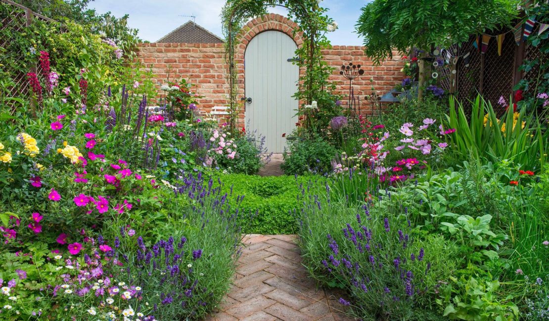 a small garden with overflowing flower beds and a brick path leading to a pretty gate at the end