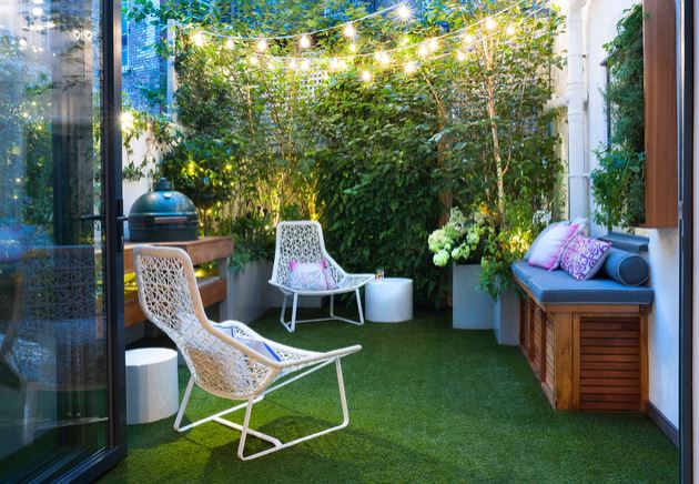 a tiny terrace with plants as fencing, string lights, slimine chairs, a storage bench and artificial grass