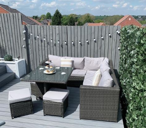 a wicker corner sofa with two footstool seats and a glass topped coffee tabe. String lights hang on the fence behind.