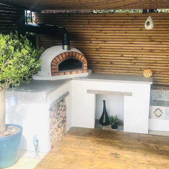 an outdoor kitchen area with a log stack and wood-fired pizza oven on a stone counter