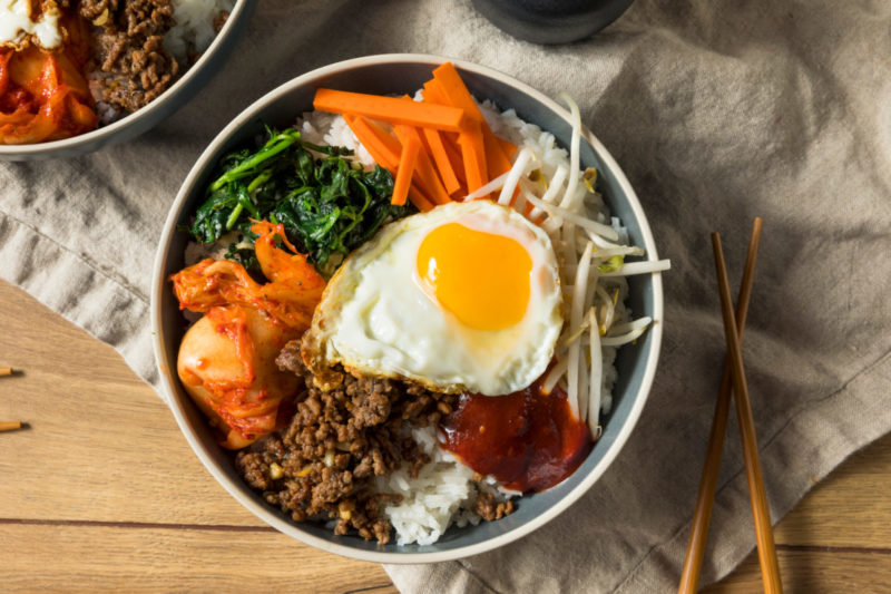 a bowl of bibimbap with fresh kitchen veggies, rice and a fried egg on top