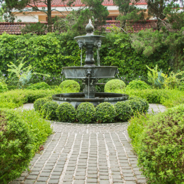 a paved path with a fountain and spherical topiary bushes in the middle
