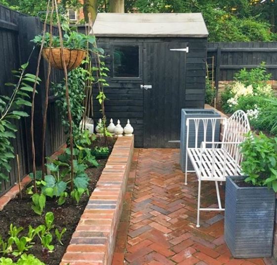 a little shed at the end of a red brick garden path with a raised vegetable patch
