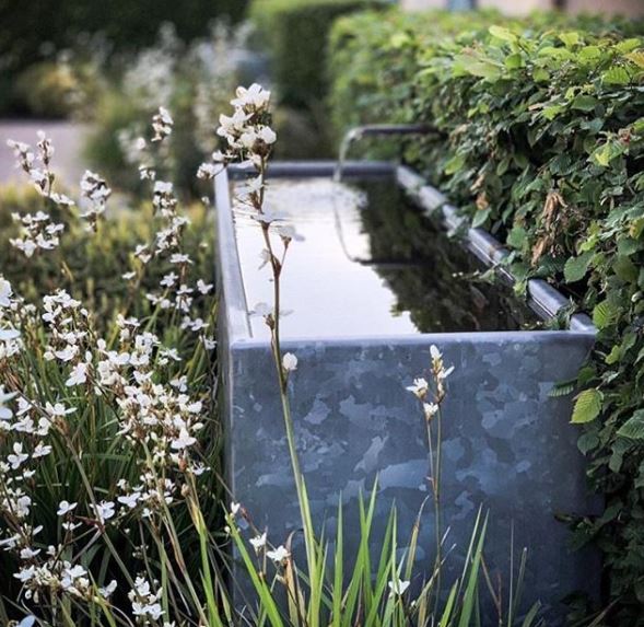 a metal trough nestled in a flower bed with a spout of water gently flowing into it