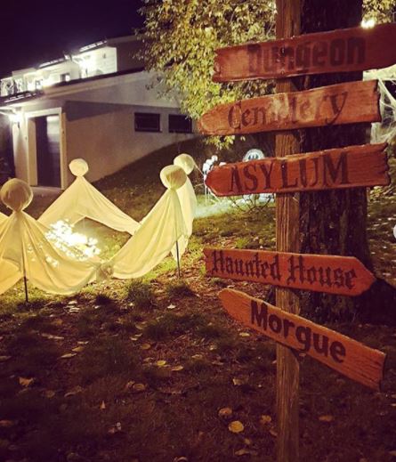 22 Halloween Garden Ideas: Fun & Low-cost Spooky Decorations 17 a wooden directions sign pointing to spooky locations, on a lawn with illuminated ghosts made from sheets