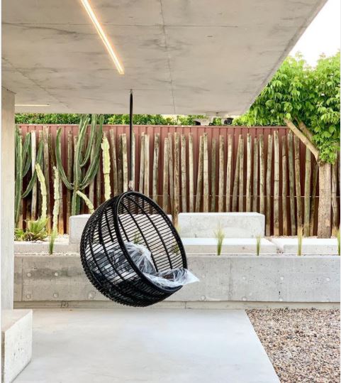 a black wicker garden swing chair hanging from a concrete roof, above a concrete floor, with a wooden fence and spiky green plants in the background