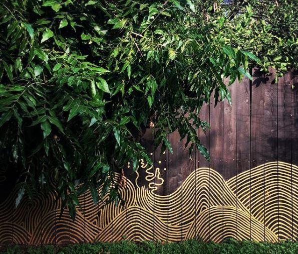29 Unique Garden Mural Ideas for Outdoor Walls & Fences 15 a brown fence partially covered by a hedge, with a line painting of ocean waves at the bottom
