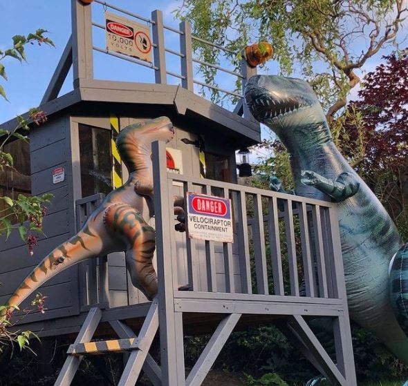 a playhouse on a platform, decorated with fencing and keep out signs to resemble a security cabin. Two inflatable dinosaurs block the entrance