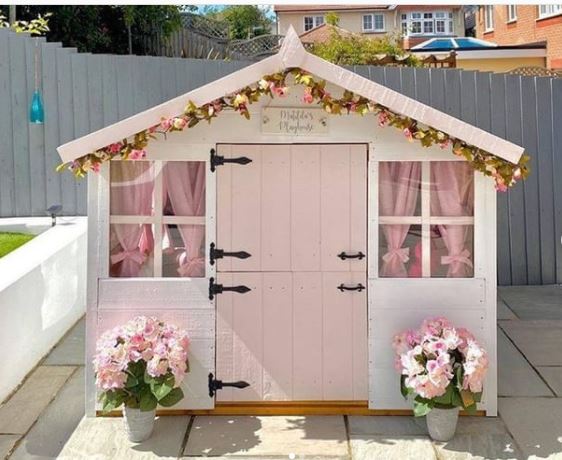 a pretty playhouse with pink curtains, a pink door and pink flowers outside