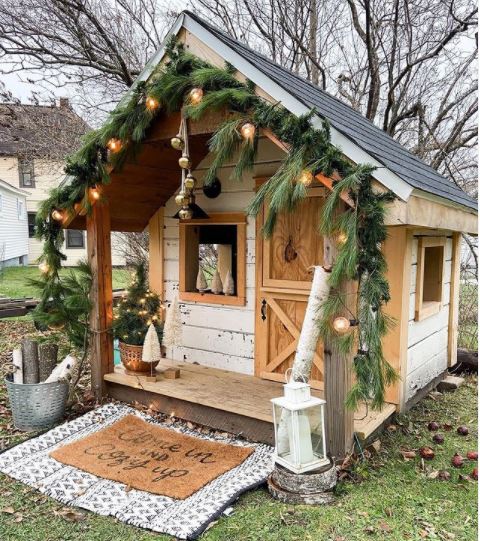 scandinavian-inspired garden playhouse ideas with wreaths of foliage, string lights, hygge style door mat and rustic decorations