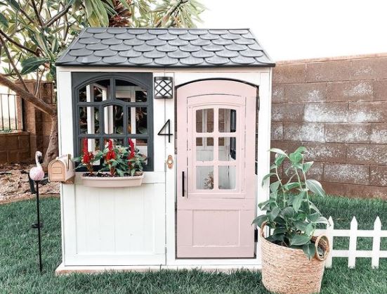 a grown-up playhouse for kids with a pastel pink door, picket fence and window box with flowers