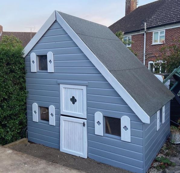 a wooden playhouse with an asymmetrical pitched roof, painted blue with a pretty white door and matching shutters