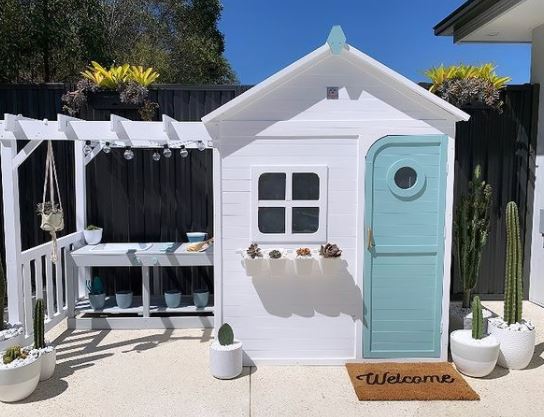 a bright white playhouse with a cheerful blue door. On the outside there is a window box, welcome mat and tiny pergola