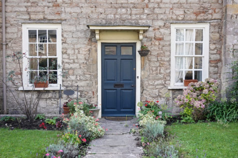 the front of a stone house with a blue front door, mowed lawn and neat flowers along the path