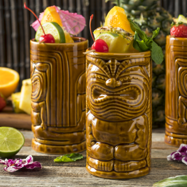 three novelty ceramic tiki mugs shaped like wood carvings are filled with exotic fruit drinks
