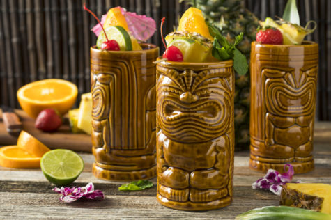 three novelty ceramic tiki mugs shaped like wood carvings are filled with exotic fruit drinks