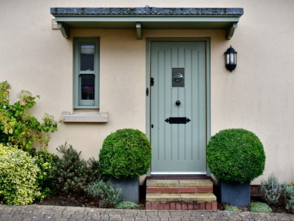 the entrance to a home with neat box shrubs either side of a grey-green door. There is an exterior light on the wall.
