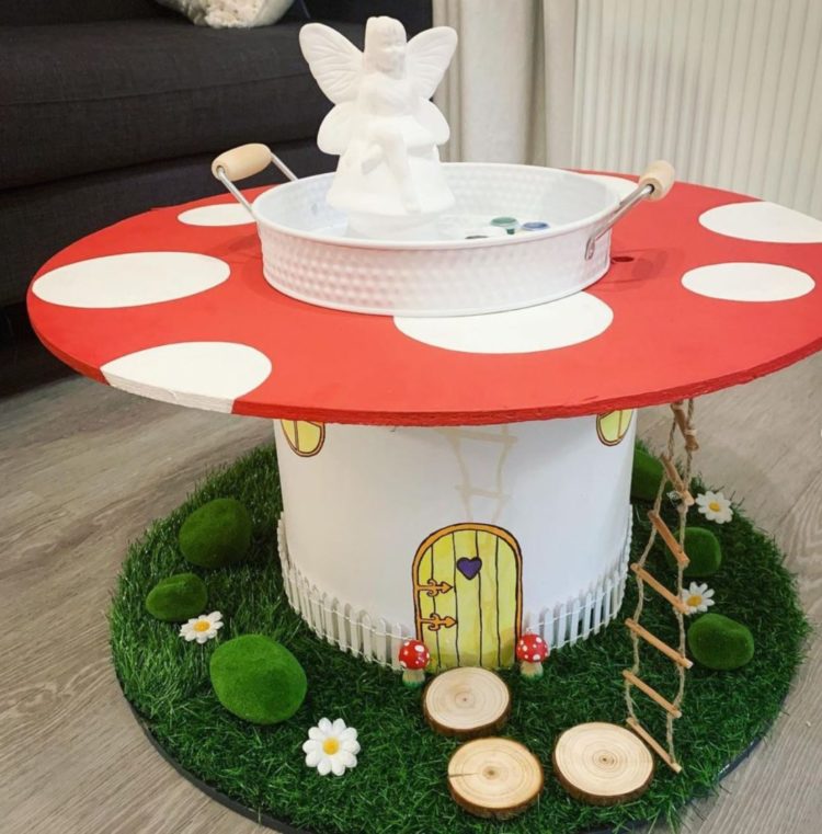 a giant cable spool has been painted to resemble a toadstool, with a miniature door, ladder and foliage