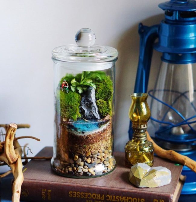 a small glass jar with sand, blue resin, green moss and a tiny figurine inside to resemble a lakeside landscape