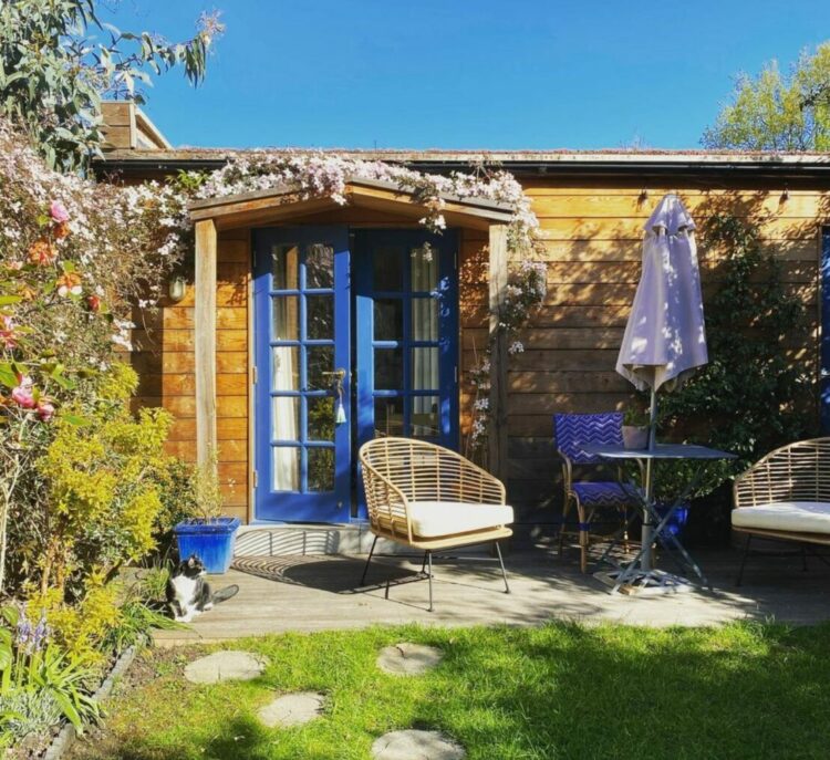 a wooden cabin with deep blue door can barely be seen behind patio furniture and a bushy clematis plant