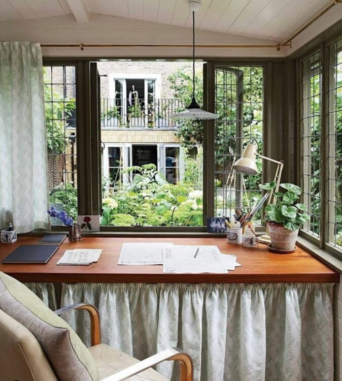a view looking over the desk of a garden office outside, through mullioned windows dressed with pale green curtains